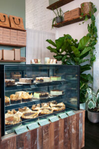 Cheesecake display counter with reclaimed timber finishes and lush greenery inside Better Burnt café.