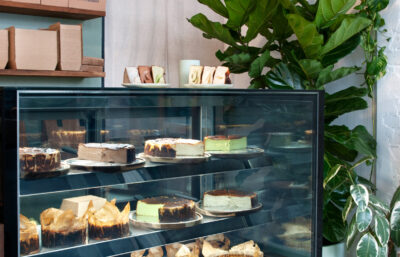 Cheesecake display counter with reclaimed timber finishes and lush greenery inside Better Burnt café.