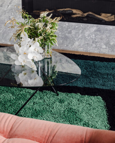 Glass coffee table with white orchids and greenery on textured green rug in Collingwood apartment.