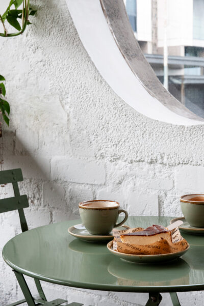 Coffee cups and cheesecake on a green café table against a textured white brick wall at Better Burnt Hawthorn.