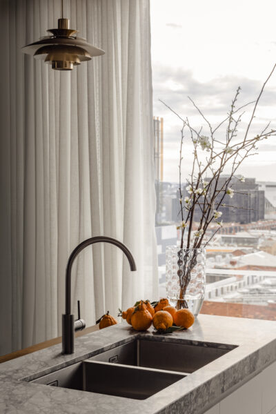 Marble kitchen counter with sink, pendant light, and vase of branches overlooking Melbourne skyline.