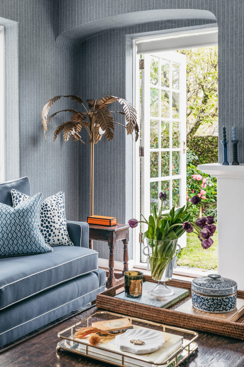 Blue living room with patterned cushions, gold palm lamp, and garden outlook.
