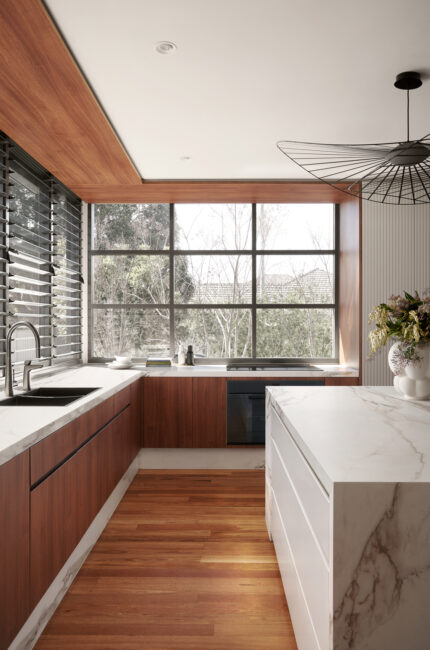 Light-filled kitchen with timber joinery, marble island, and large windows framing nature.