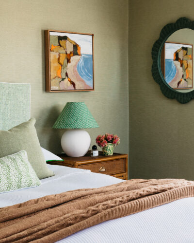 Bedside table with green lamp, coastal painting, and textured green wallpaper.
