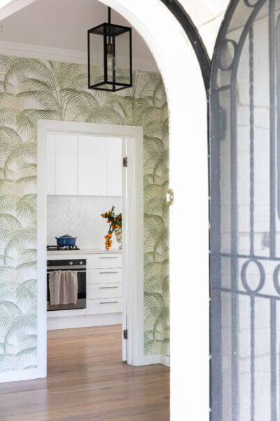 View through arched doorway to kitchen with palm wallpaper and white cabinetry.